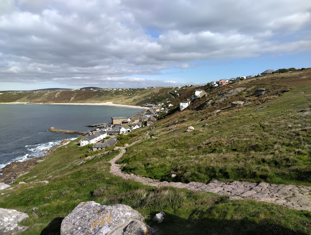 A photo of a seaside town of white-walled buildings nestled above a still blue cove. A cobbled path on a grassy hill winds down from the viewer towards the town. A stony pier juts out into the bay. Behind the town, more steep coastline and hills sweep into the distance.